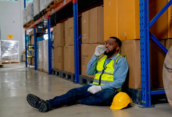 African American man sit on floor and lean to boxes on shelve then drink water to relax after work in workplace area.