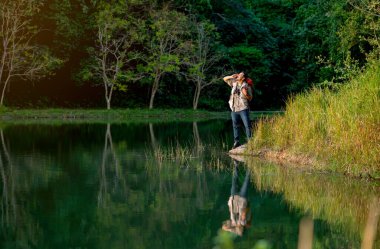 Çantalı ve dürbünlü Asyalı adam ya da diğer aksesuarlı seyahat için çığlık at ya da bağır ve gölün kenarında dur. Kamp yaparken gölgesinin yansımasıyla milli parka git..