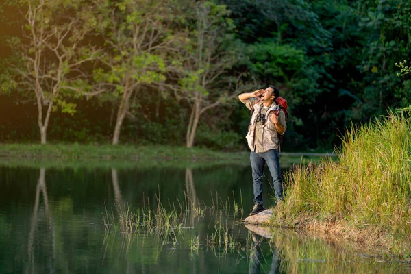 stock image Asian man with bagpack and binoculars or other accessories for travel action of shout or bawl and stand near lake during camping and travel in national park.