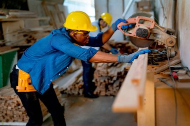 Afrikalı Amerikalı marangoz, fabrika işyerindeki keresteleri kesmek için elektrikli testereyi kullanmak için dikkatli bak ve iş arkadaşı hala arkada çalışıyor..