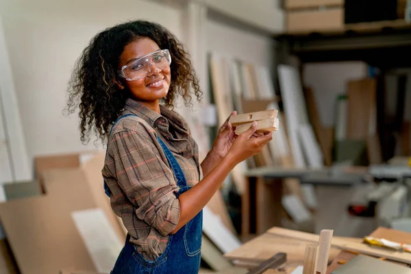 Smart Asian carpenter woman with African hair style hold piece of wood and look at camera with smiling in concept of happiness during work support good quality of product.