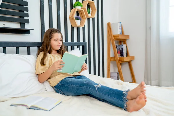 Little Caucasian girl enjoy to read book and learn on bed with day light in bedroom alone in concept of happiness activities at home.