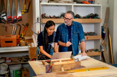 Asyalı ahşap işçisi kadın, müşterinin sipariş detaylarını kontrol etmek için dizüstü bilgisayar kullanıyor ve beyaz kocasıyla internetten ahşap ürün satışı hakkında konuşuyor..