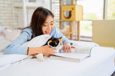 Asain bir kadın yatakta uzanıyor ve yanında uzanan çok renkli kedi için kitap okuyor. Gün ışığı ile birlikte. İyi ilişki kavramı ve evcil hayvan ve hayvan arasındaki bağ kavramı..