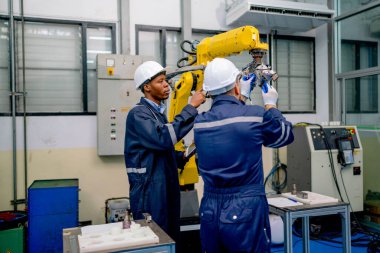 Afrikalı Amerikalı teknisyen veya mühendis robotik kolu işaret ediyor ve fabrika işyerindeki makine sorununu çözmek için iş arkadaşına rehberlik ediyor.