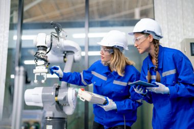 İki Kafkasyalı profesyonel mühendis veya teknisyen kadın işçi kontrol ve tablet yardımıyla fabrika işyerinin yanında duran robotik kol makinesini kontrol ve bakım için.