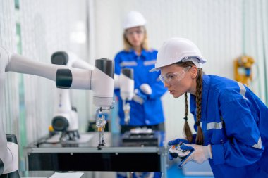Profesyonel mühendis veya teknisyen robotik kolla çalışmak için denetleyici kullanır ve fabrika işyerindeki makinenin bakımıyla birlikte arka planda iş arkadaşının önünde durur..