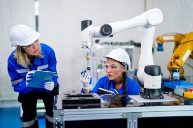 İki profesyonel teknisyen veya mühendis kadının ön görüntüsü oturur ve fabrika işyerindeki robotik makine ile sistem kontrol ve ipad yardımcı olur.
