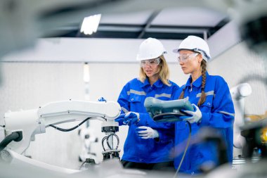 Güzel mühendis veya teknisyen işçiler, fabrika işyerindeki robotik kolu kontrol etmek ve kontrol etmek ve fabrikadaki diğer makine veya kanalları kullanarak görüntülemek için birlikte çalışırlar..