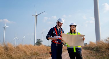 Kafkasyalı mühendis ve kadın çizim kağıdı kullanarak tartışır ve mavi gökyüzü olan rüzgar türbininin önünde dururlar..