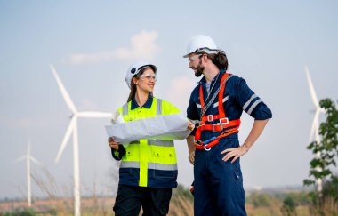 Kafkasyalı mühendis, iş arkadaşıyla kağıt çizerken mavi gökyüzü olan rüzgar türbini ya da yel değirmeninin önünde durur..