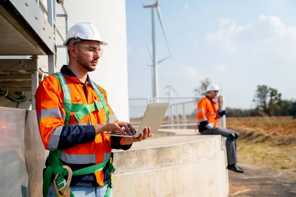 One of engineer or technician worker stand with using laptop to work near his co-worker at base of windmill or wind turbine with blue sky and day light.
