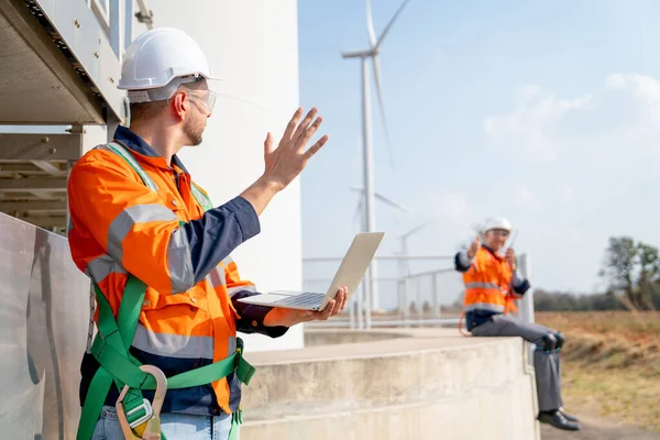 Caucasian engineer or technician worker hold laptop and act as greeting to his co-worker, they stay on base of big windmill or wind turbine with blue sky.