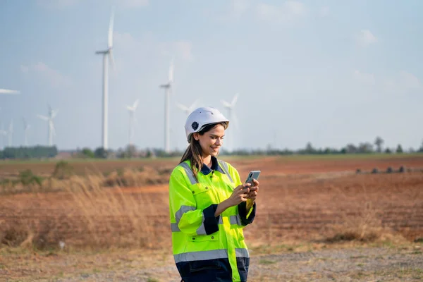 Pretty engineer or technician woman with safety protection equipment stand with using mobile phone and stand in front of windmill cluster in the field.