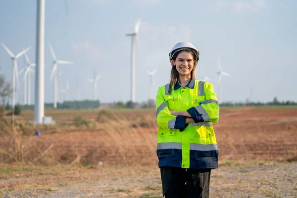 Pretty engineer woman with safety protection equipment stand with arm-crossed and look at camera in front of windmill cluster in the field.