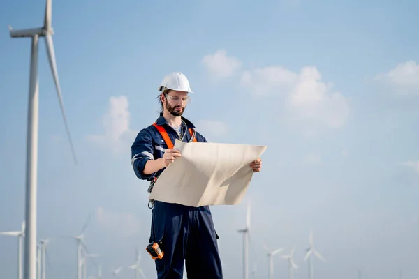 Caucasian engineer or technician man stand with holding drawiing paper and look to the paper and stand in front of windmill or wind turbine cluster and blue sky.