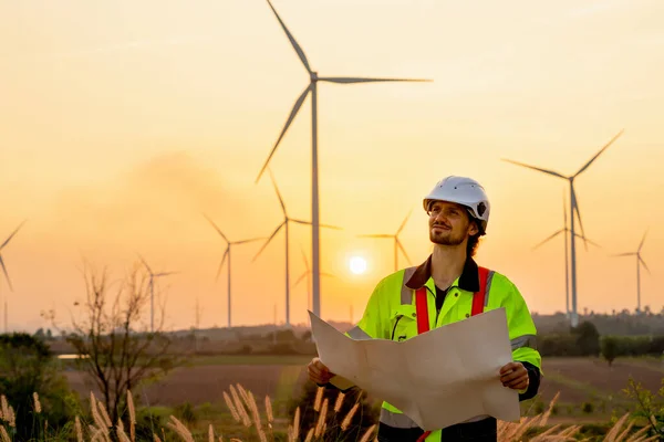 Caucasian engineer or technician man stand with holding drawiing paper and look to left side and stand in front of windmill or wind turbine cluster with sunset and warm light.