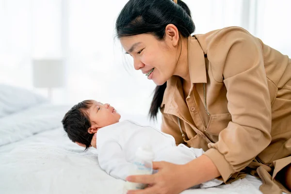 Asian mother hold newborn baby who look at her mother also stay on bed in bedroom of their house with day light.