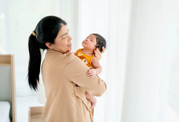 アジアの母親は 彼女の胸の上に眠って新生児を保持し 昼光と白いカーテンで窓の前に立つと 彼女は赤ちゃんを世話する幸せそうに見えます — ストック写真