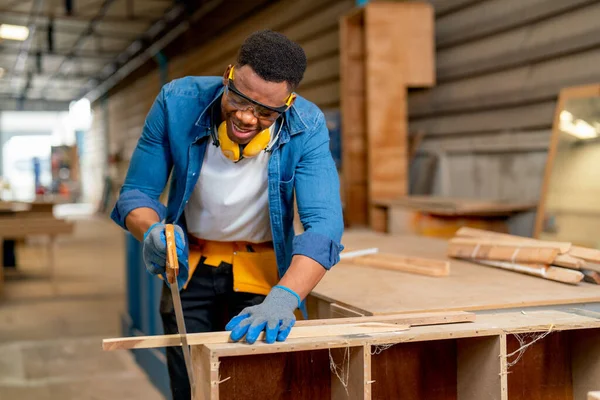 African American man as carpenter use saw to cut timber in factory or workplace with happiness and express smiling during work.