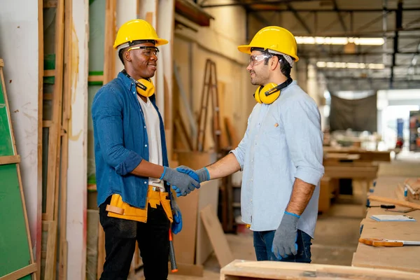 African American and Caucasian carpenter man shake hands together with smiling after success in wood work project in factory workplace.