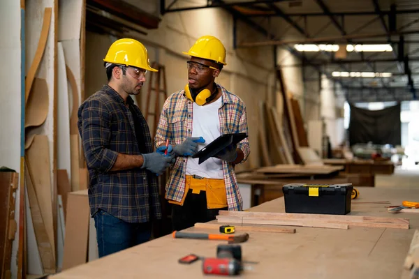 African American and Caucasian carpenter man discuss together about wood work using tablet in factory workplace.