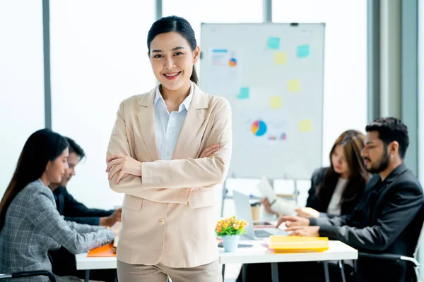Portrait of Asian business woman wear bright suit stand with arm-crossed and look at camera show confidence in front of other co-workers discuss on the back in office.