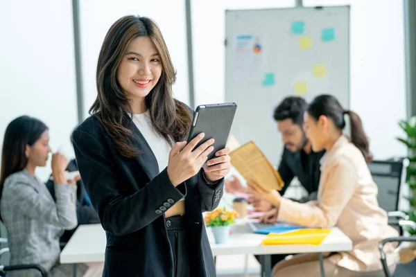 Portrait of Asian business woman hold tablet and stand with look at camera in front of other co-workers discuss on the back in office.