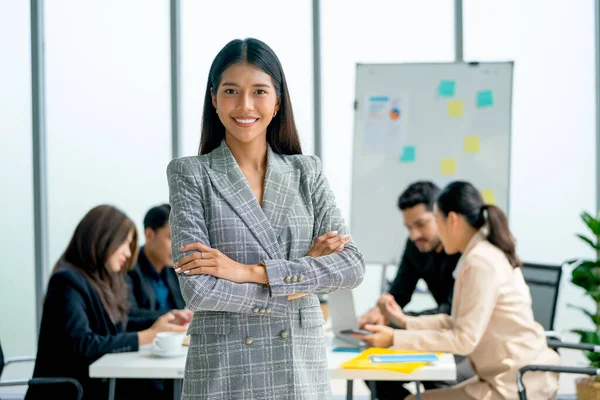 Portrait of Asian business woman stand with arm-crossed and look at camera show confidence in front of other co-workers discuss on the back in office.