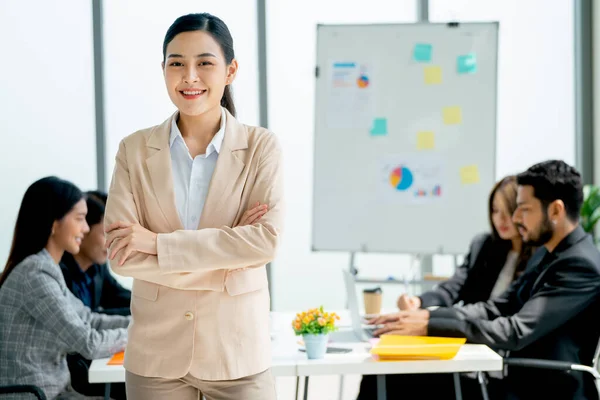 Portrait of Asian business woman wear bright suit stand with arm-crossed and look at camera show confidence in front of other co-workers discuss on the back in office.