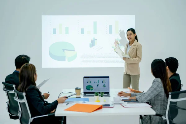 Pretty Asian business woman stand in front of meeting room and present on white wall that projection by the projector to coworkers and manager.