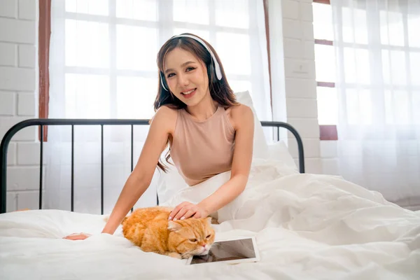 Beautiful Asian woman with headphone sit and pat orange cat that lie over tablet on bed and she also look at camera with smiling.