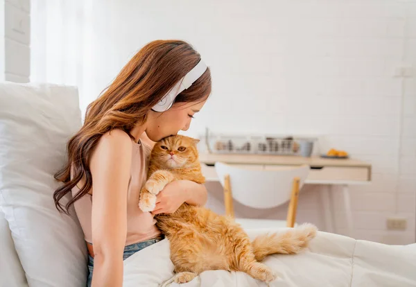 헤드폰을 주황색 고양이와 키스하는 아시아 자신의 침실에 침대에 — 스톡 사진