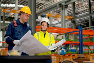 Fabrika işçileri, iş yerinde makine ve ürün rafları olan kağıt ya da plan kullanarak projeyi tartışıyor..