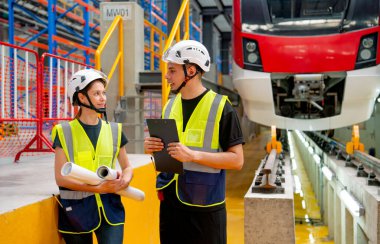 Çalışan fabrika işçileri ve kadın çalışanlar, iş yerindeki elektrikli tren projesiyle ilgili bir döküman platformunun arka planında..