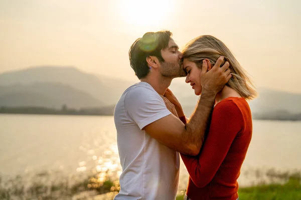 Close up Caucasian man kiss on forehead of beautiful woman with sunset light near lake and look romantic for couple love stay together.