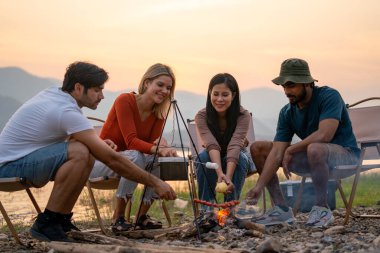 Çok ırklı bir grup erkek ve kadın arkadaş partide yemek yemekten ve ateşle çevrili oturmaktan hoşlanıyorlar ve göl ve gün batımı yakınlarındaki halk parkında bir araya gelmekten mutlu görünüyorlar..