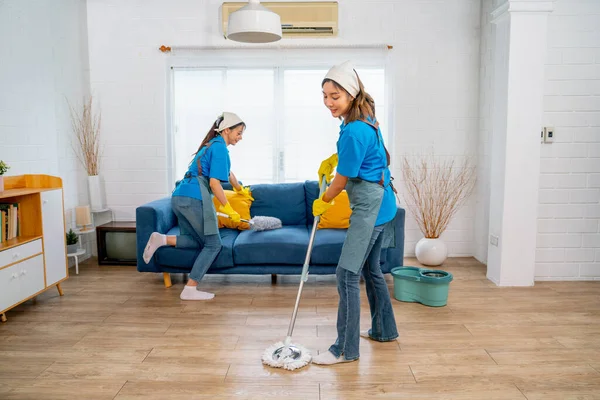 Asian housemaid or housekeeper team help to clean living room together with one use mop clean floor and other set pillow on sofa in the back.