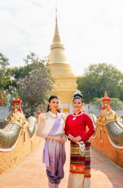 Tayland geleneksel elbiseli iki Asyalı kadının dikey görüntüsü, güzel sanat ve mimari ile antik pagoda yolunda yan yana duruyor, aynı zamanda gülümsüyor ve kameraya bakıyorlar..