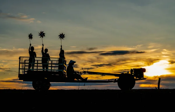 Silhouette of children hold small windmill and stand on E-taen farm tractor with driver to go back home in early morning with warm sunrise light.