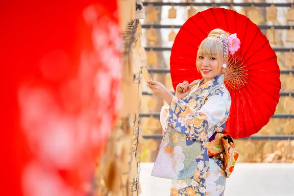 美しい女性の肖像画は日本のスタイルのドレスを着て ランタンの列の前に木製の旗に魅力的な間に赤い傘を保持します — ストック写真