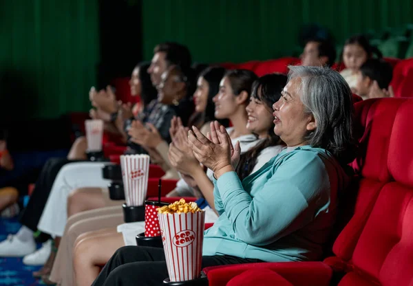 Side view of multiethnic and multigeneration people clap hands during watch movie together in cinema theater and they look happy in concept of enjoy entertainment on holiday.