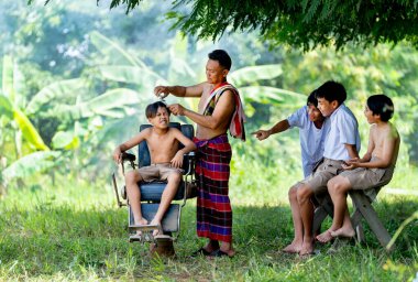 Genç oğlanların yan görünüşü eğlenceli ve parodiyi ifade eder Asyalı adam tarafından geleneksel berber olarak kesilen arkadaşlarını büyük bir ağacın altında..