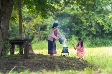 Asyalı kadın anne gibi görünüyor. Pirinç tarlasının yanında pilav pişirmeye hazırlanıyor. Kızı da mutluluktan yanında oturuyor..