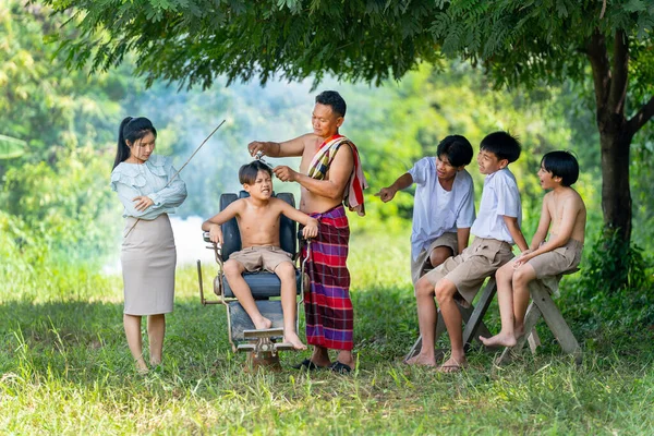 Asian man work with haircut to boy on chair with the teacher and friends that look fun and parody the boy and they stay outdoor under big tree.