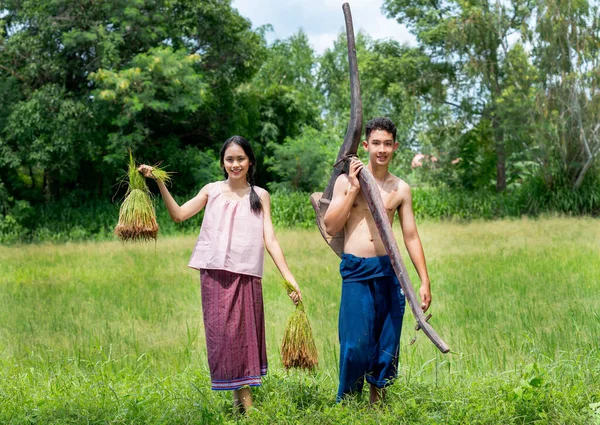 Portrait of young Asian boy and girl with carry plow and rice seedlings stand on way near rice field with day light.