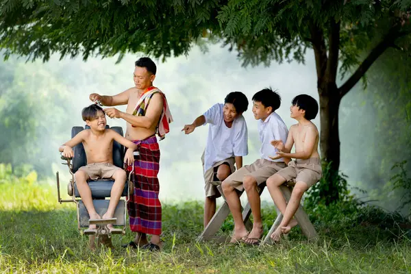 男孩子们在大树下嬉笑着模仿他们的朋友 这位朋友被亚洲男人当作传统理发师理发 图库图片