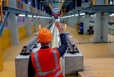 Güvenlik üniformalı teknisyen arkası elektrik veya metro trenlerine vücut sinyali veriyor ve fabrika işyerindeki demiryolu raylarının önünde duruyor..