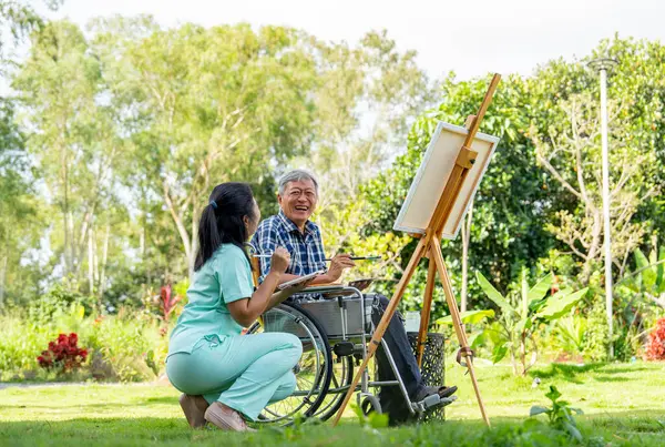 Back of Asian nurse woman sit near senior man on wheelchair enjoy to paint the painting outdoor in the garden and they look happiness with laughing together.