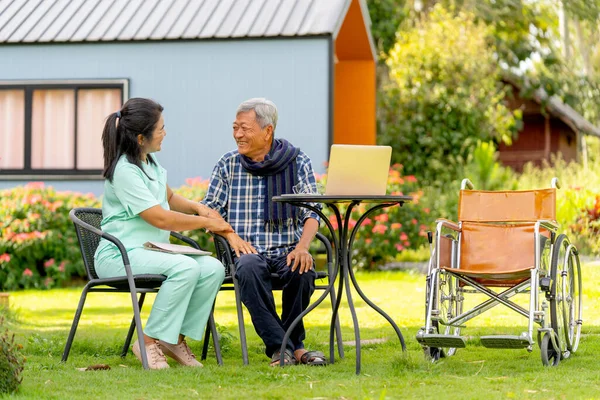 Asian senior man and nurse relax outdoor in the garden with laptop is on the table and wheelchair is set beside.